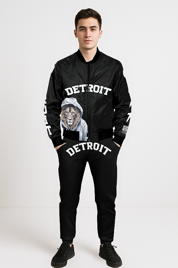 A man standing against a light background, wearing a matching black Detroit-themed outfit. The black bomber jacket features the word 'DETROIT' in bold white letters across the chest and an illustration of a lion wearing a hoodie on the left side. The jacket sleeves also have 'DETROIT' printed vertically in white. The matching black joggers display 'DETROIT' in bold white letters across the front waistband. The man is wearing black sneakers and has his hands in his pockets.