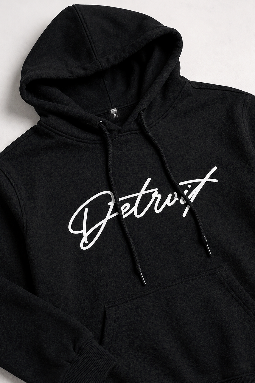 Flat lay detail of a black hoodie angled left on a clean white background, featuring a white cursive “Detroit” graphic across the chest, visible drawstrings, hood structure, and front pocket stitching with soft studio lighting.