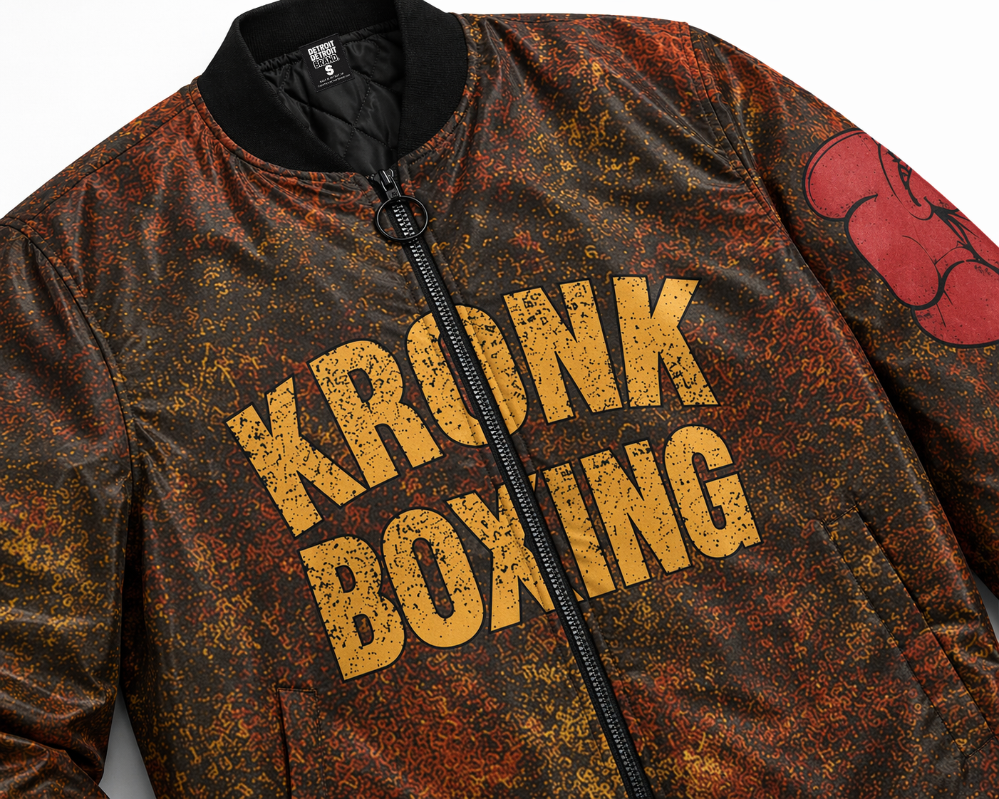 Close-up angled flat lay of KRONK Boxing bomber jacket with distressed rust texture, bold yellow lettering, zipper detail, and boxing glove graphic on sleeve