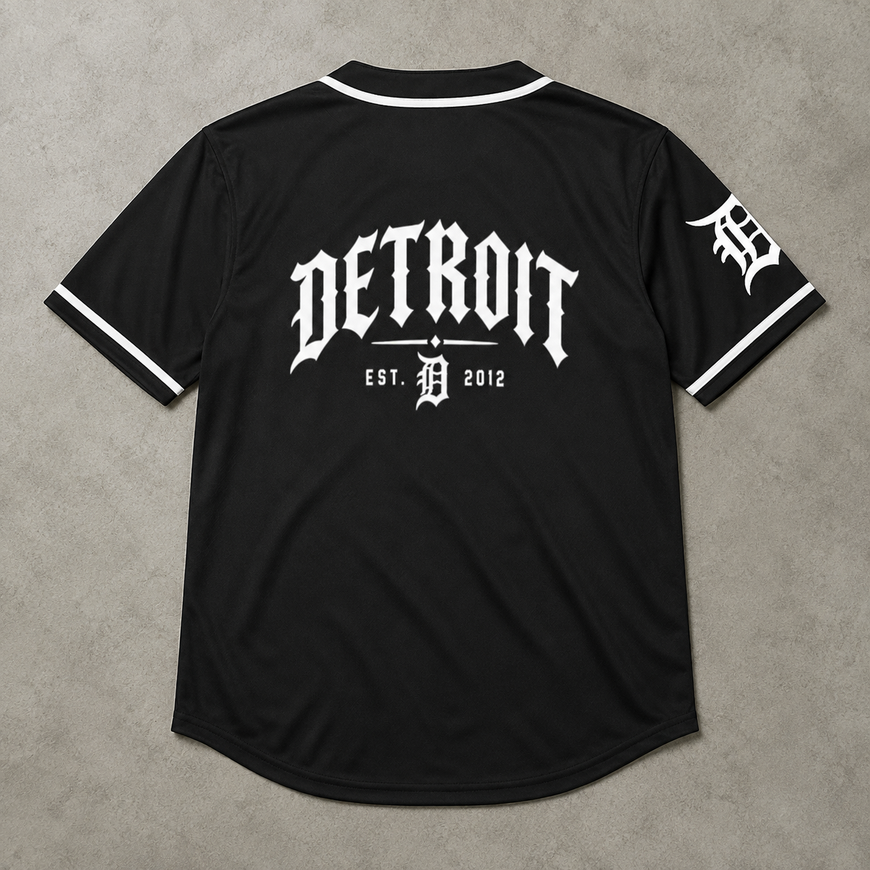 Black Detroit Tigers baseball-style jersey shown from the back, featuring a large white 'DETROIT' logo and 'EST. 2012' with a stylized 'D' in the center, plus a white 'D' logo on the sleeve, laid flat on a gray textured background.