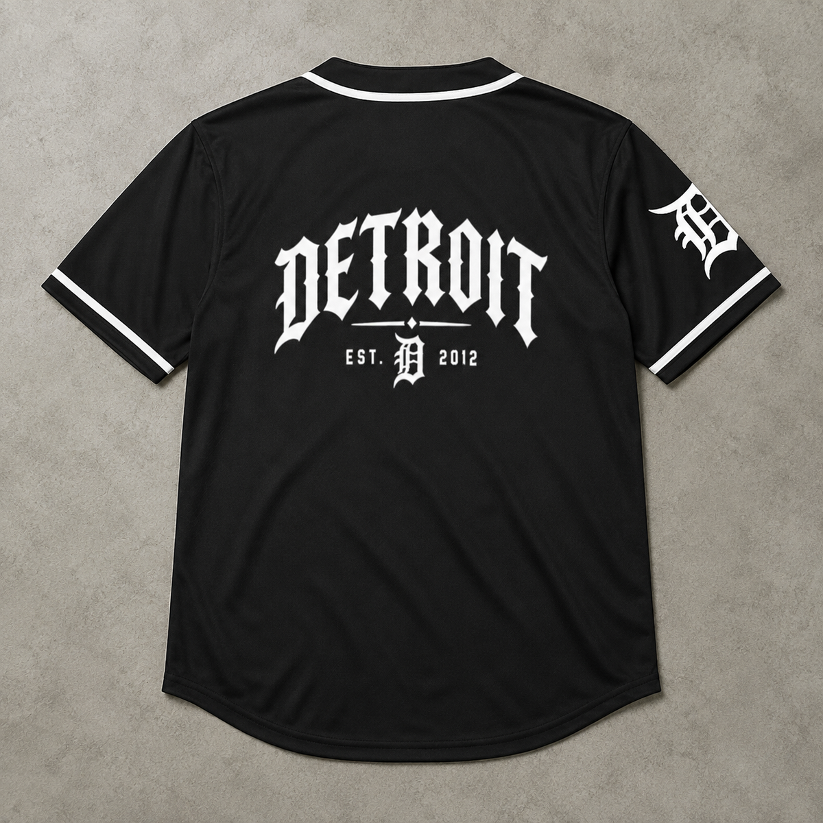 Black Detroit Tigers baseball-style jersey shown from the back, featuring a large white 'DETROIT' logo and 'EST. 2012' with a stylized 'D' in the center, plus a white 'D' logo on the sleeve, laid flat on a gray textured background.