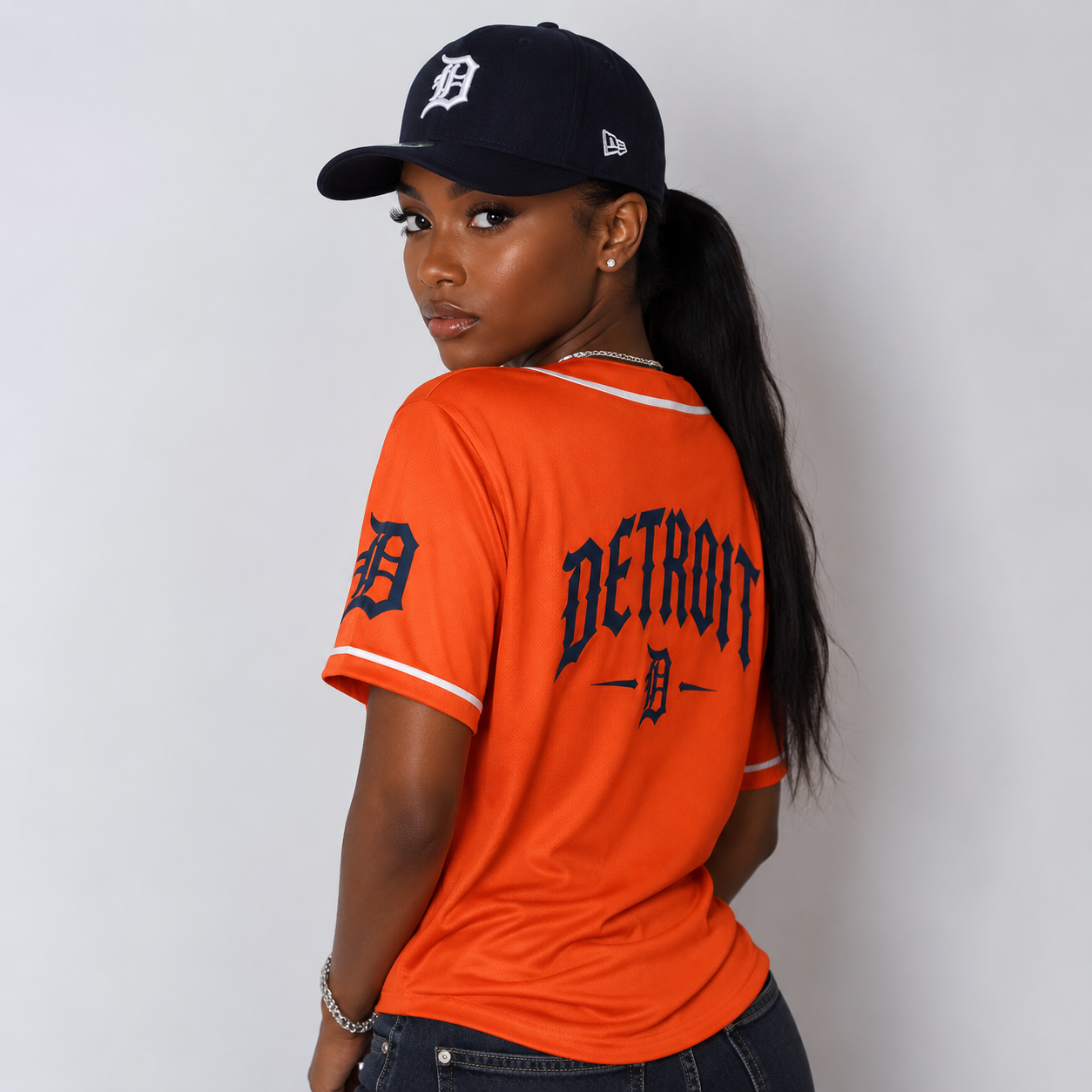 A woman wearing a bright orange Detroit Tigers baseball jersey with navy blue lettering and white trim, paired with a navy cap, photographed against a plain light gray wall.