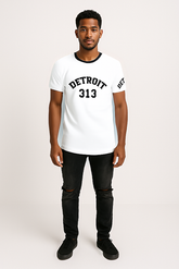 A man standing against a light-colored background, wearing a white t-shirt with a black crew neckline. The front of the t-shirt features the word &