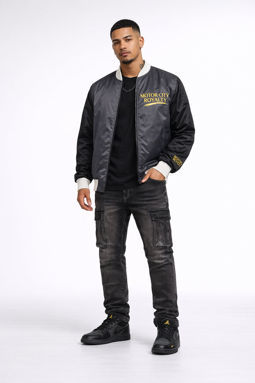 Man wearing a black bomber jacket with 'Motor City Royalty' text, black pants, and black shoes on a white background