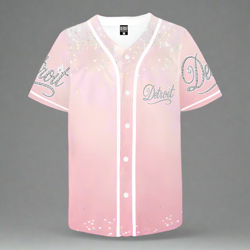 Pink short-sleeve baseball jersey displayed flat against a gray background. The jersey features white buttons and white piping along the front placket, collar, and sleeves. The word 'Detroit' is written in a silver glittery script font on the left chest and both sleeves. The upper part of the jersey has a sparkling confetti-like design, adding a glamorous touch