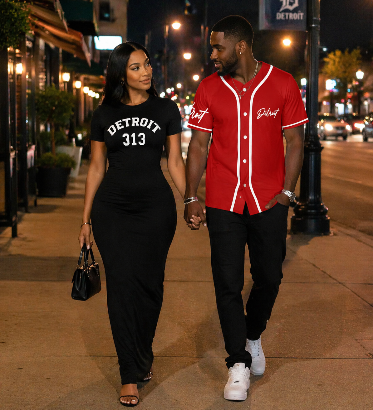 Couple walking on a city street at night wearing Detroit streetwear, woman in fitted black Detroit 313 maxi dress and man in red Detroit baseball jersey, urban date night lifestyle scene