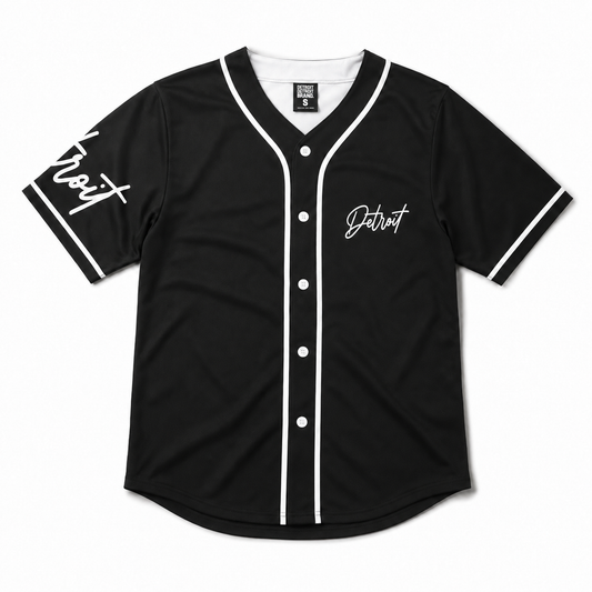 Front flat lay of a black Detroit baseball jersey with white piping, white buttons, white interior neckline, and Detroit script graphics on a white studio background.