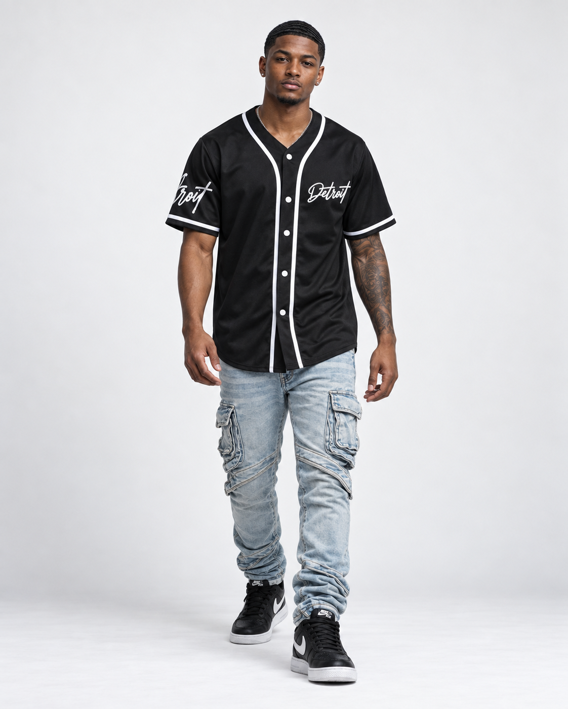 Brown skin male model wearing a black Detroit baseball jersey with white piping, light wash cargo jeans, and black sneakers on a white studio background.