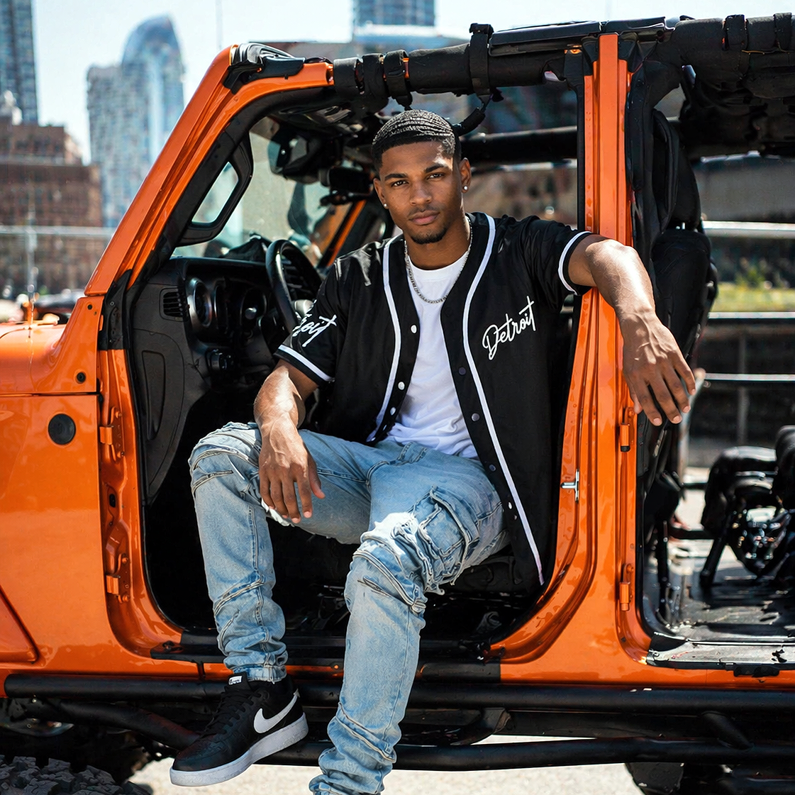Man wearing black Detroit baseball jersey unbuttoned sitting inside orange Jeep Wrangler Rubicon, urban streetwear lifestyle outfit