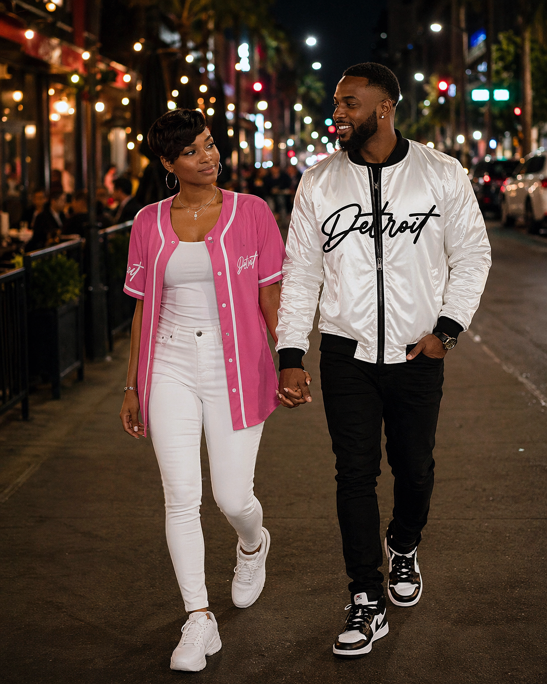 Couple walking at night with man wearing white and black Detroit bomber jacket and woman in pink Detroit jersey, date night city street scene