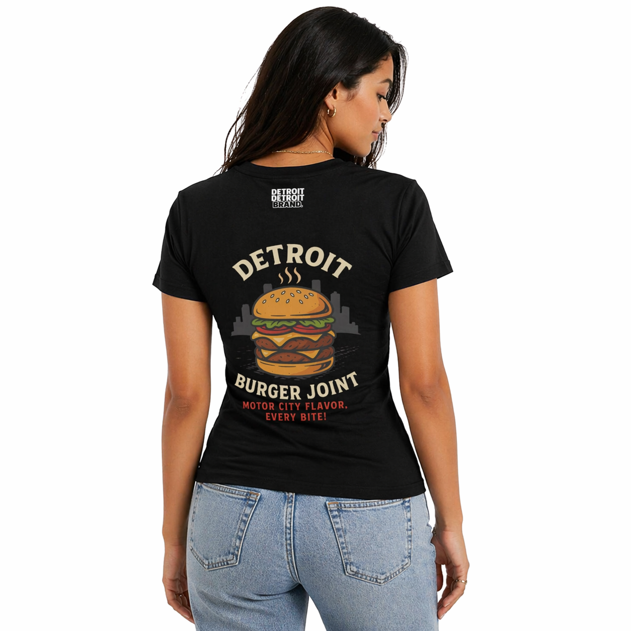 Back view of female model wearing a fitted black Detroit Burger Joint graphic t-shirt with large burger design and “Motor City Flavor Every Bite” text, paired with light blue jeans on a white studio background