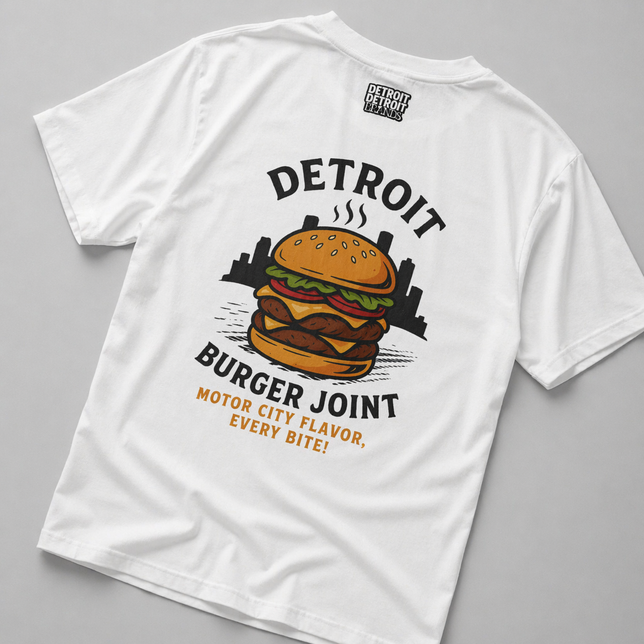 Flat lay detail shot of white Detroit Burger Joint t-shirt back showing burger graphic, Detroit skyline, and “Motor City Flavor, Every Bite!” text
