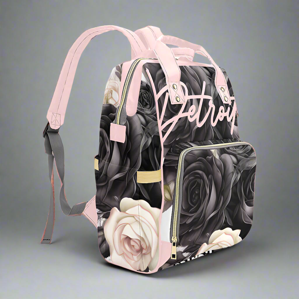 Stylish backpack featuring a black and gray rose pattern with soft pink accents, gold zippers, and “Detroit” written in pink script on the front pocket.