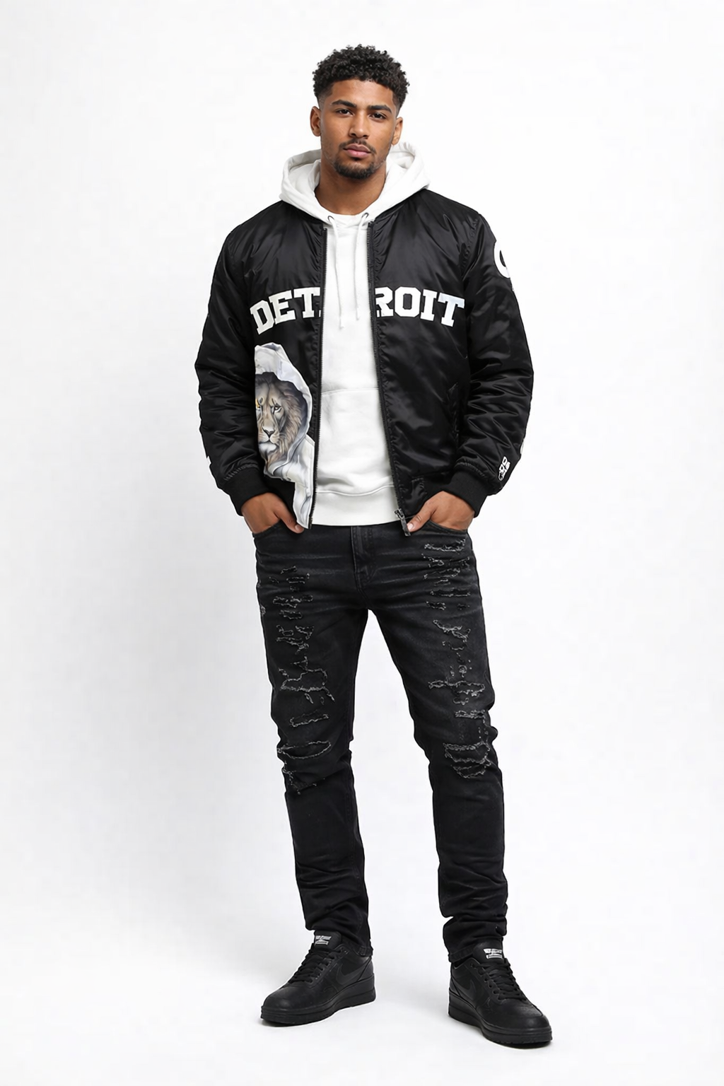 Male model wearing black Detroit lion graphic bomber jacket with white hoodie, black jeans and black sneakers, front view