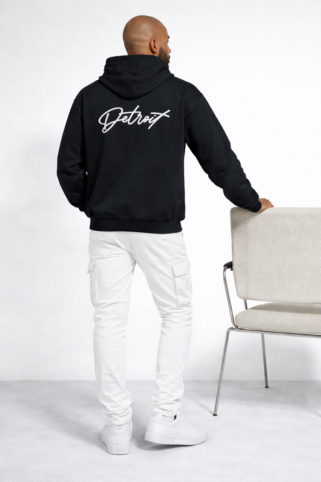 Back view of man wearing black Detroit script hoodie with large Detroit logo, styled with white pants in studio setting