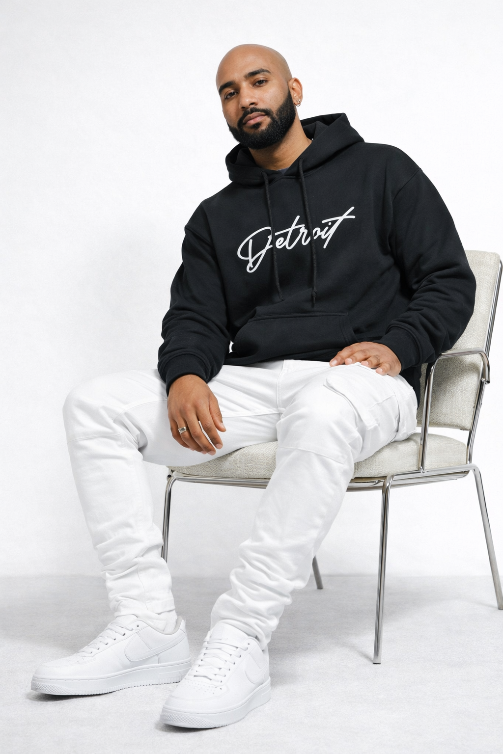 Man sitting wearing black Detroit script hoodie front view with white pants and sneakers, clean studio lifestyle look
