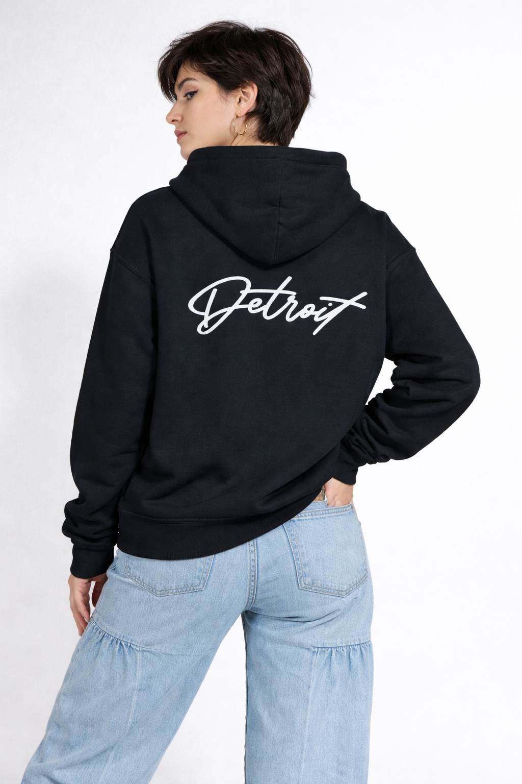 Back view of woman wearing black Detroit script hoodie showing large white Detroit logo on back, styled with light blue jeans
