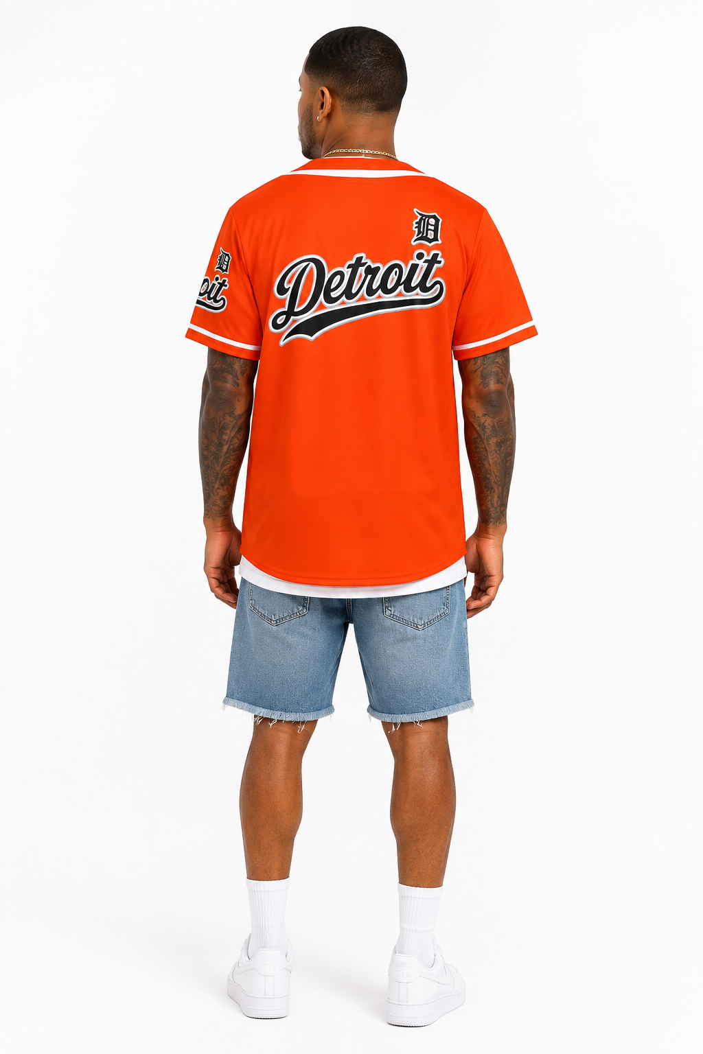 Rear view of a brown skin male model wearing the Detroit Script Orange Baseball Jersey with denim shorts and white sneakers on a clean white photography background.