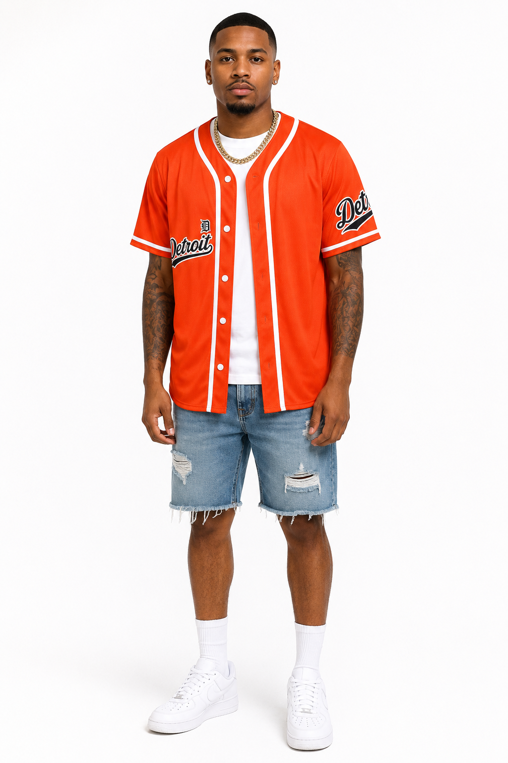 Brown skin male model wearing the Detroit Script Orange Baseball Jersey with blue denim shorts, white sneakers, white crew socks, and a gold Cuban link necklace on a white studio background.