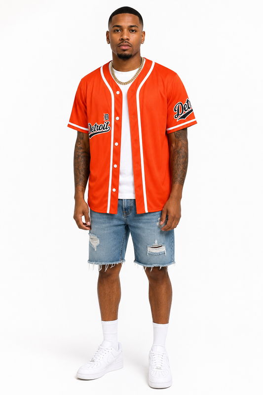Brown skin male model wearing the Detroit Script Orange Baseball Jersey with blue denim shorts, white sneakers, white crew socks, and a gold Cuban link necklace on a white studio background.