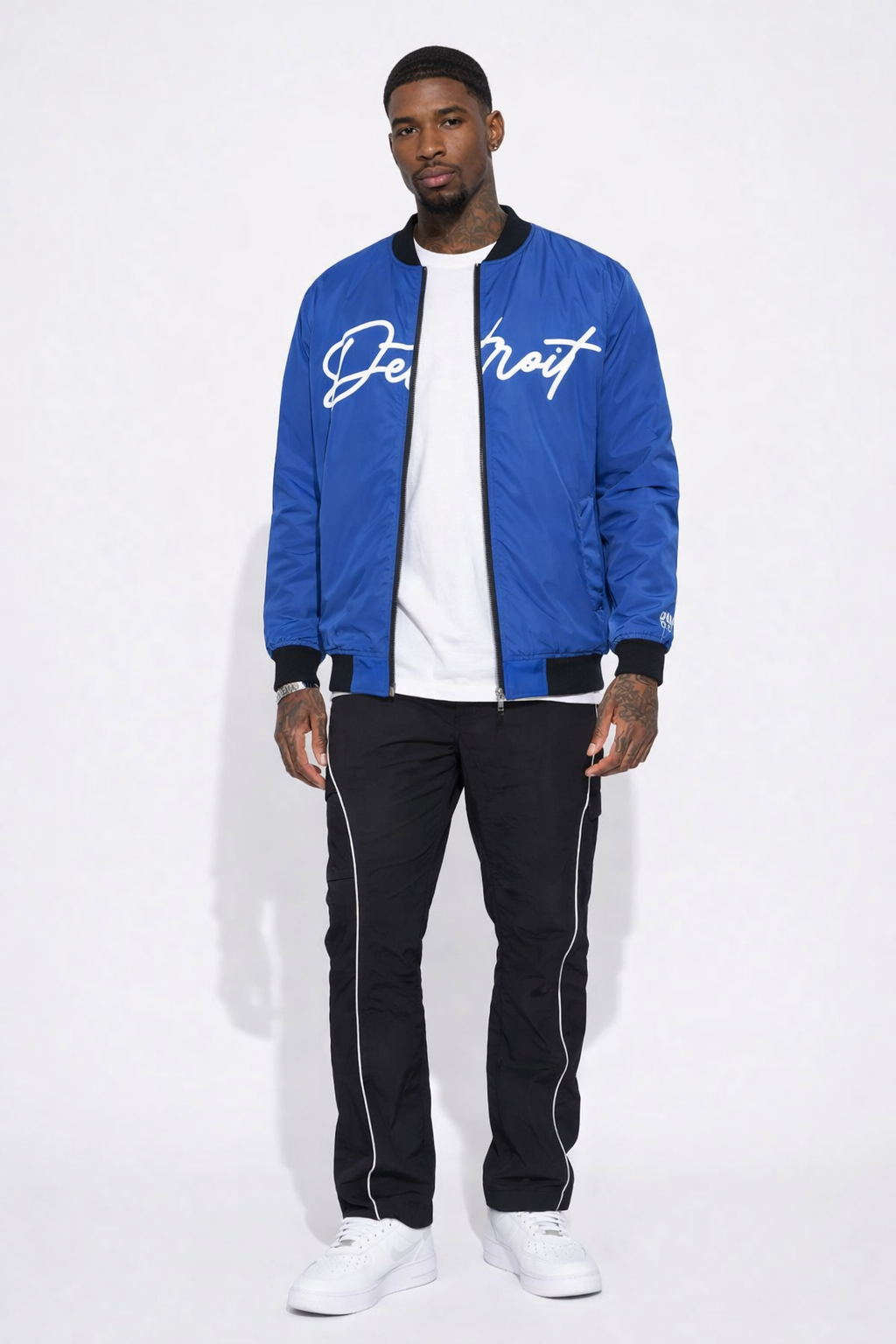 Man wearing a royal blue bomber jacket with white text, white shirt, black pants, and white sneakers on a white background