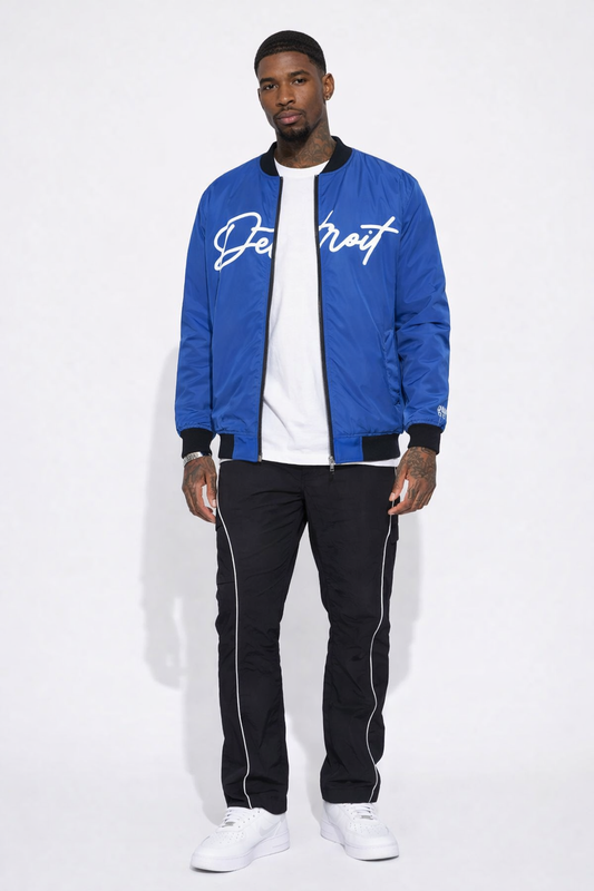 Man wearing a royal blue bomber jacket with white text, white shirt, black pants, and white sneakers on a white background