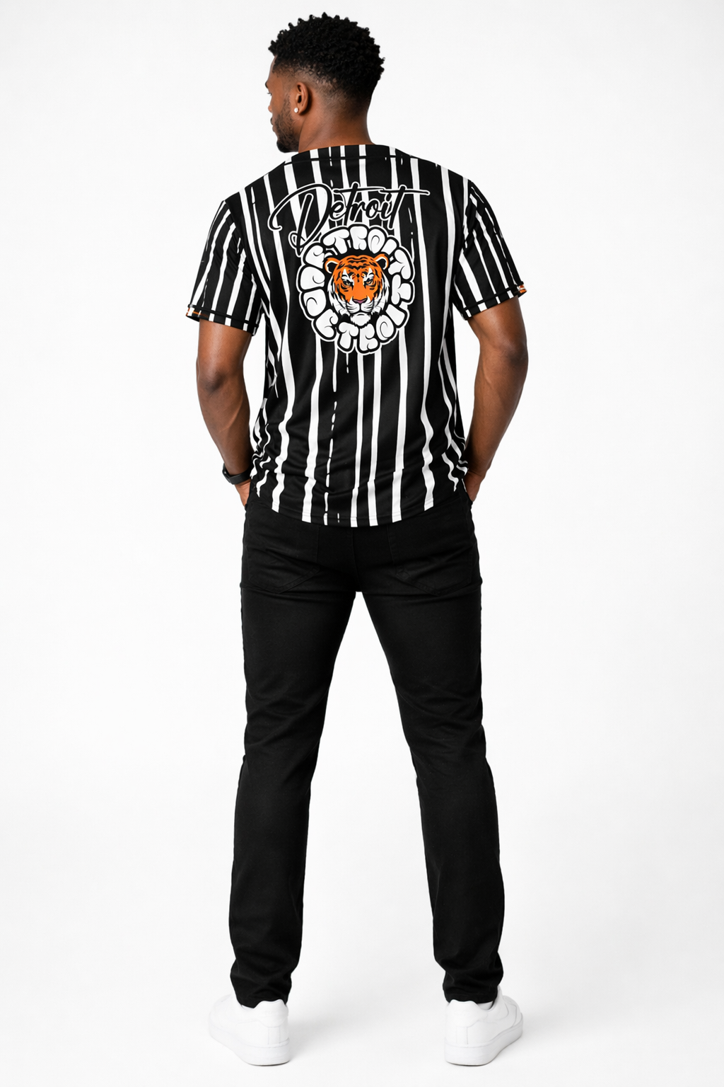 Full body back view of a male model wearing a black and white striped Detroit baseball jersey featuring a large tiger graphic and Detroit script on the back, styled with black pants and white sneakers
