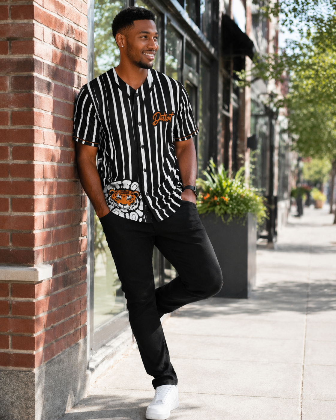 Lifestyle image of a male model wearing a black and white striped Detroit baseball jersey leaning against a brick wall in an urban street setting with natural lighting and modern storefront background