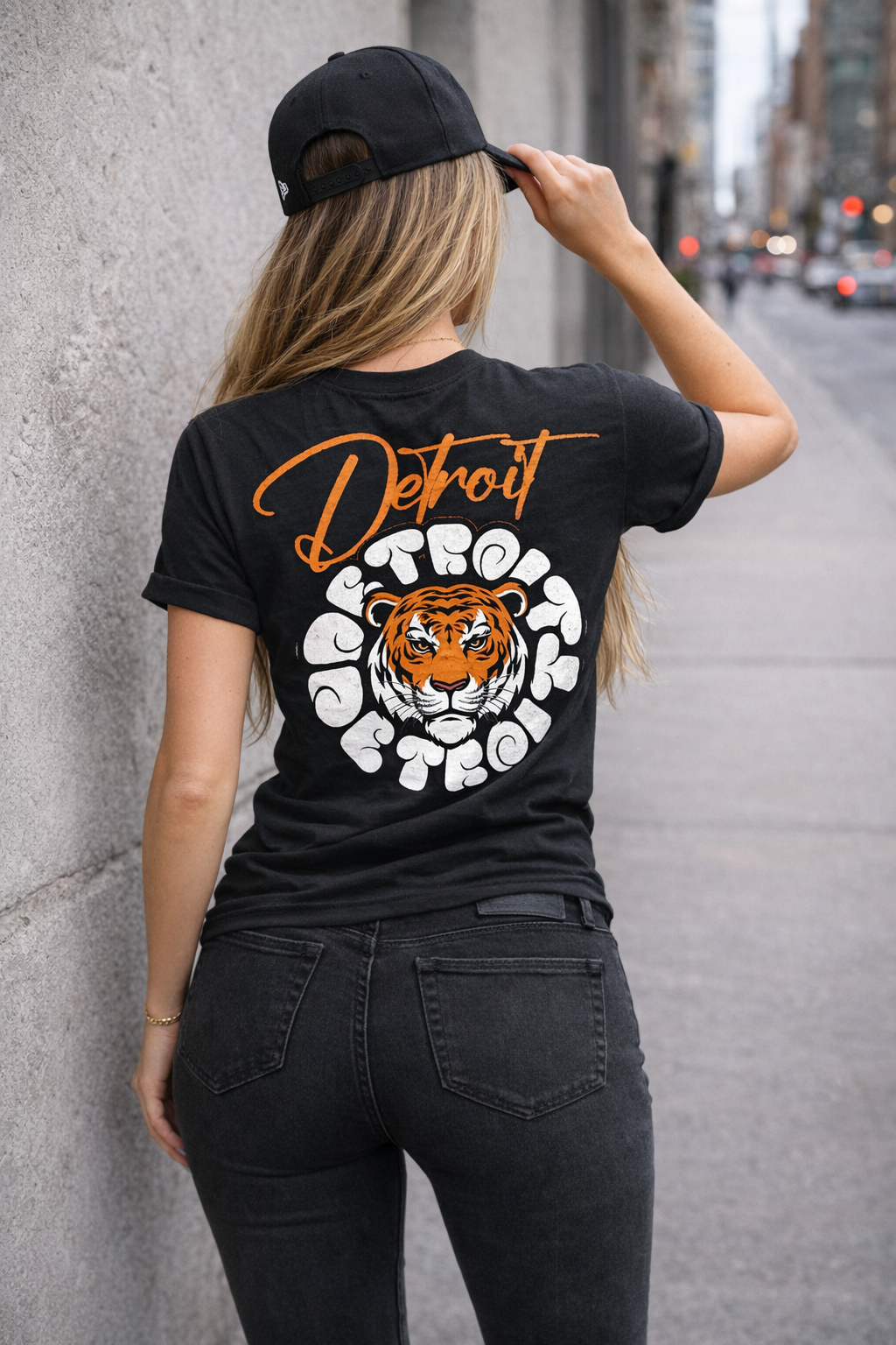 Woman wearing black Detroit t-shirt with tiger graphic back print, styled with black jeans and cap, urban streetwear lifestyle shot on city sidewalk