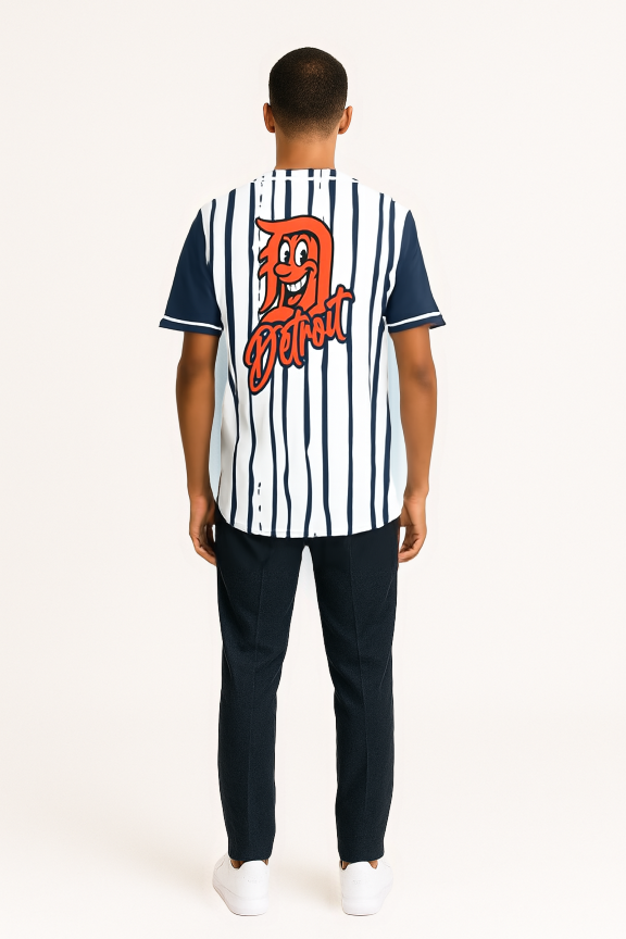 Back view of a man wearing a white baseball jersey with navy pinstripes and sleeves, featuring a large orange “D” logo and “Detroit” script graphic on the back, paired with dark pants and white sneakers, standing against a light background