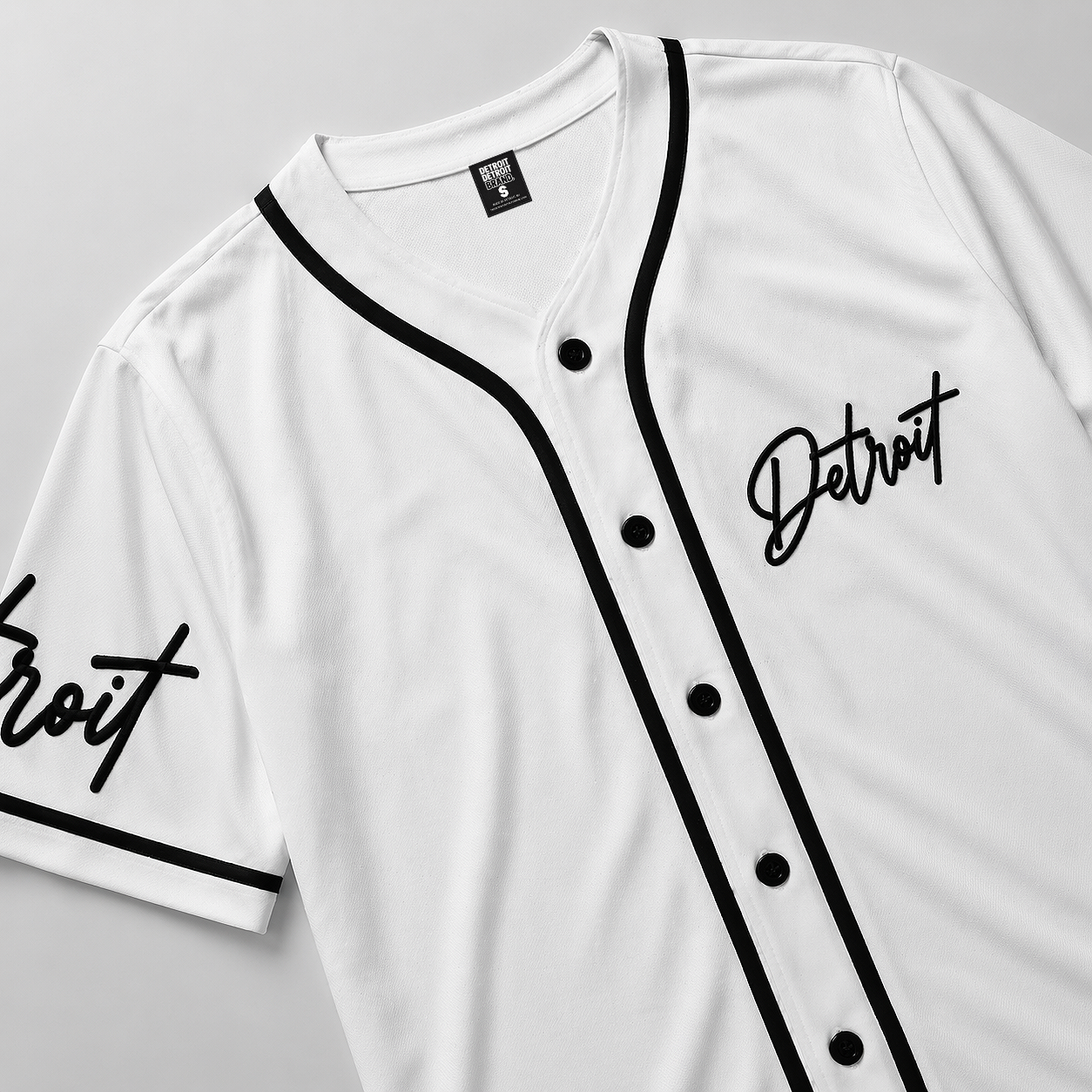 Close-up flat lay of white Detroit baseball jersey showing black piping, buttons, and script logo detail on premium fabric