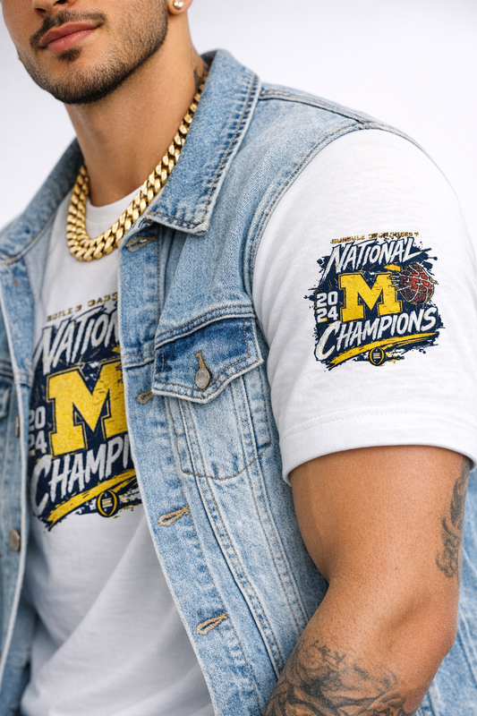 Close-up of a male model wearing a white Michigan championship t-shirt with a detailed sleeve patch, styled with a light denim jacket and thick gold chain on a clean white background.