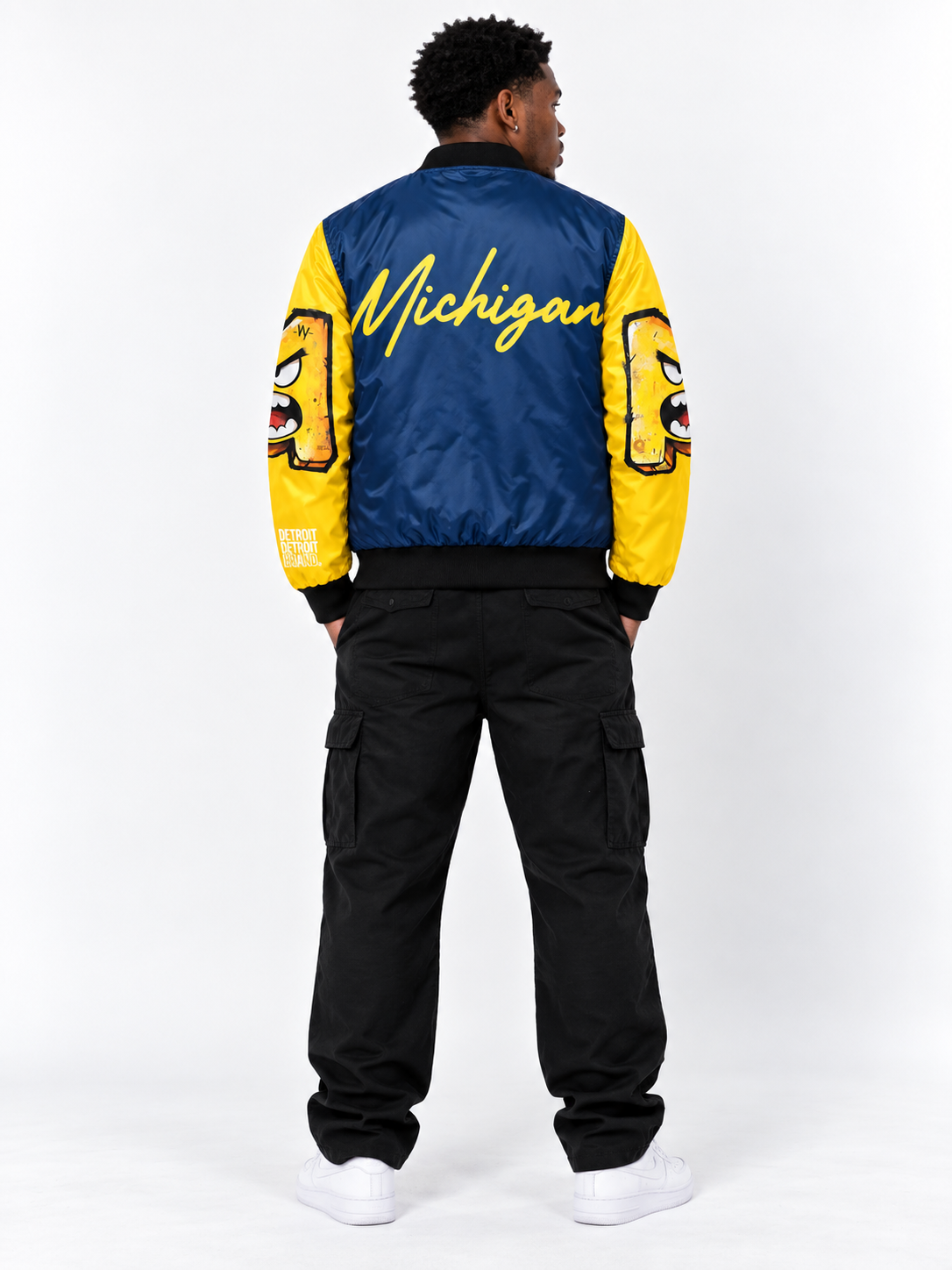 Black male model wearing the Michigan Illustrated Bomber Jacket from the back, featuring blue and maize colors, Michigan script, and illustrated sleeve graphics.