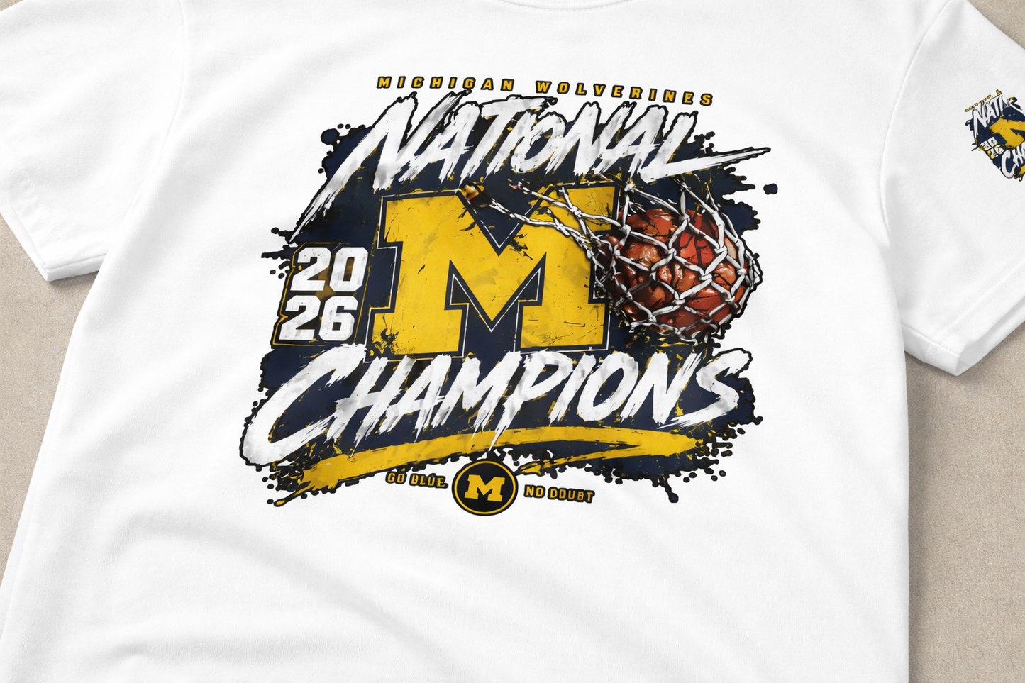 Flat lay of a white Michigan Wolverines National Champions 2026 t-shirt featuring a bold yellow “M” logo, championship text, and basketball net graphic on a clean neutral background