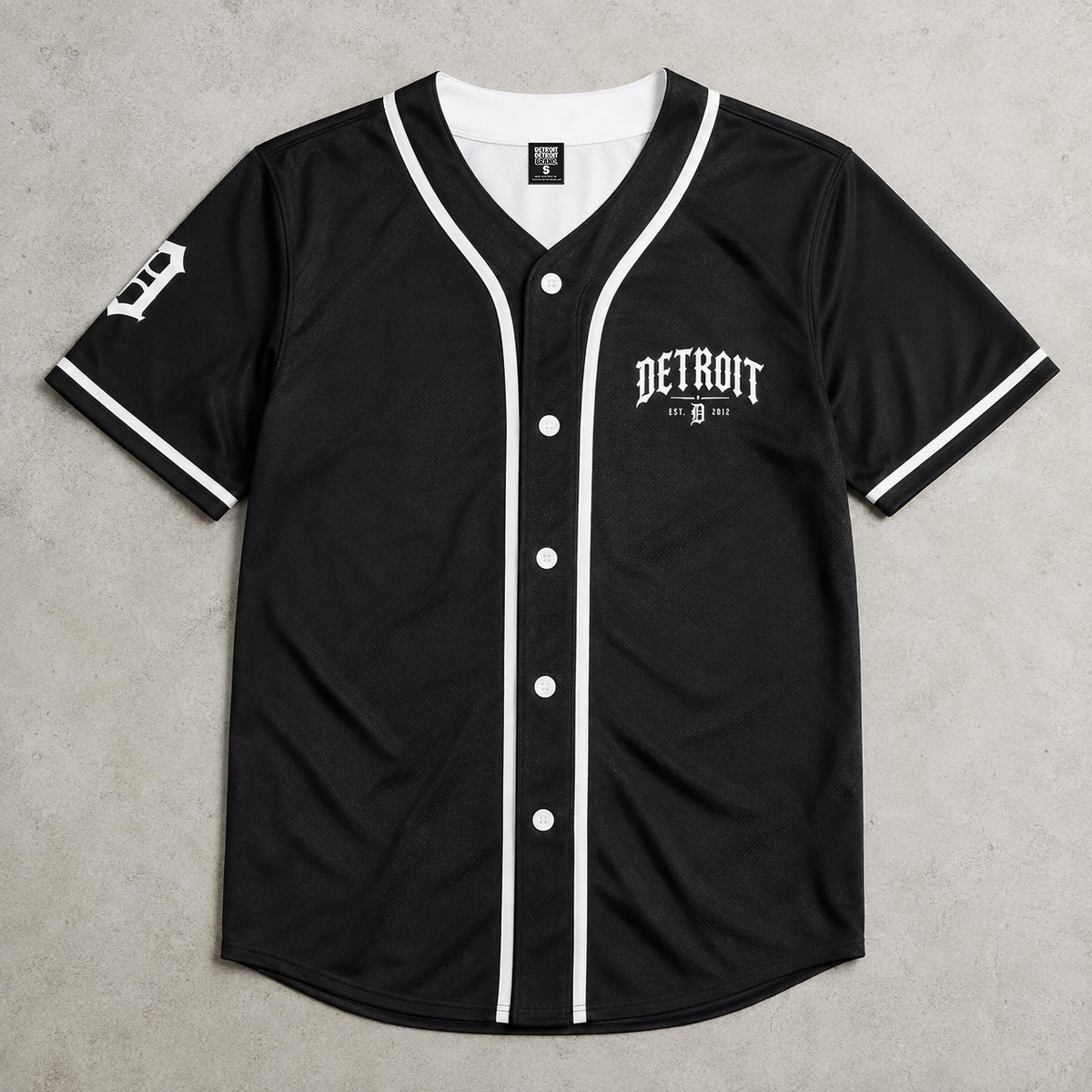 A black Detroit Tigers baseball jersey with white trim, featuring the team's 'DETROIT' logo on the left chest and a 'T' logo on the right sleeve, laid flat on a textured gray background.
