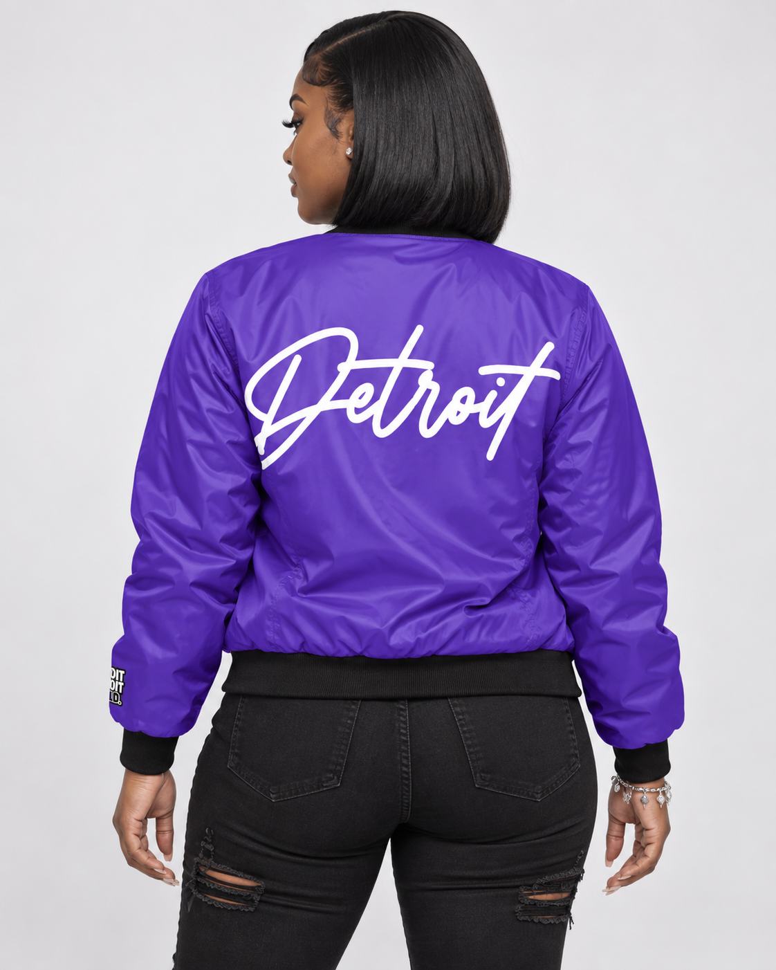 Female model wearing purple Detroit bomber jacket showing large white Detroit script on back
